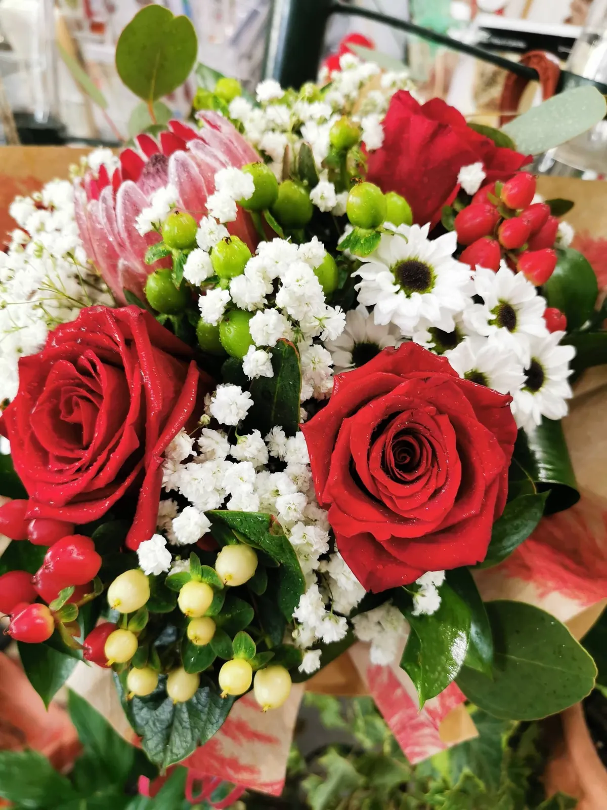 Pormenor de um arranjo de rosas vermelhas com margaridas brancas e hypericum, feito no atelier